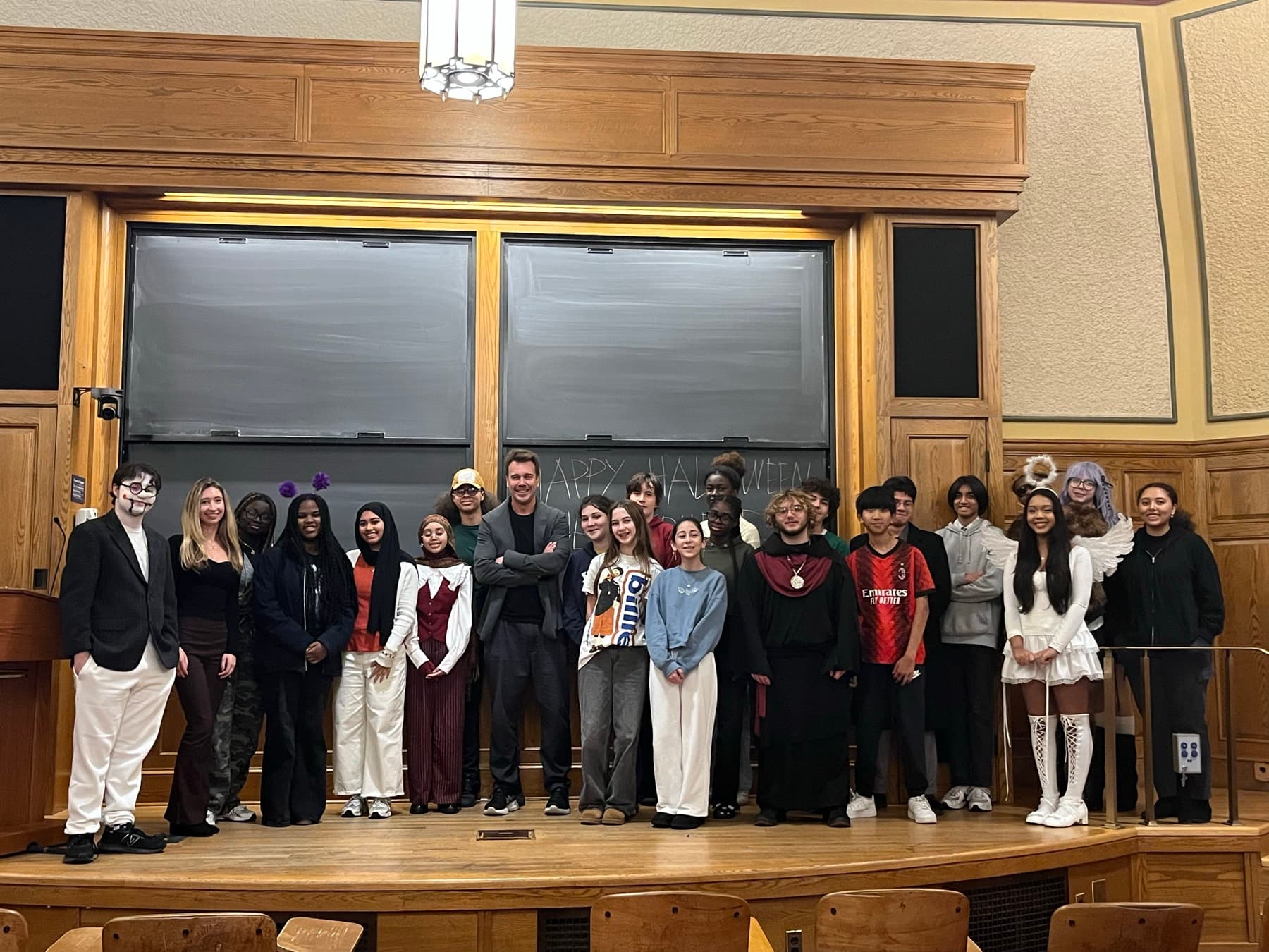 Students and their Yale teachers standing along the front of a wood-panelled Yale lecture hall, in costume, in front of a blackboard.