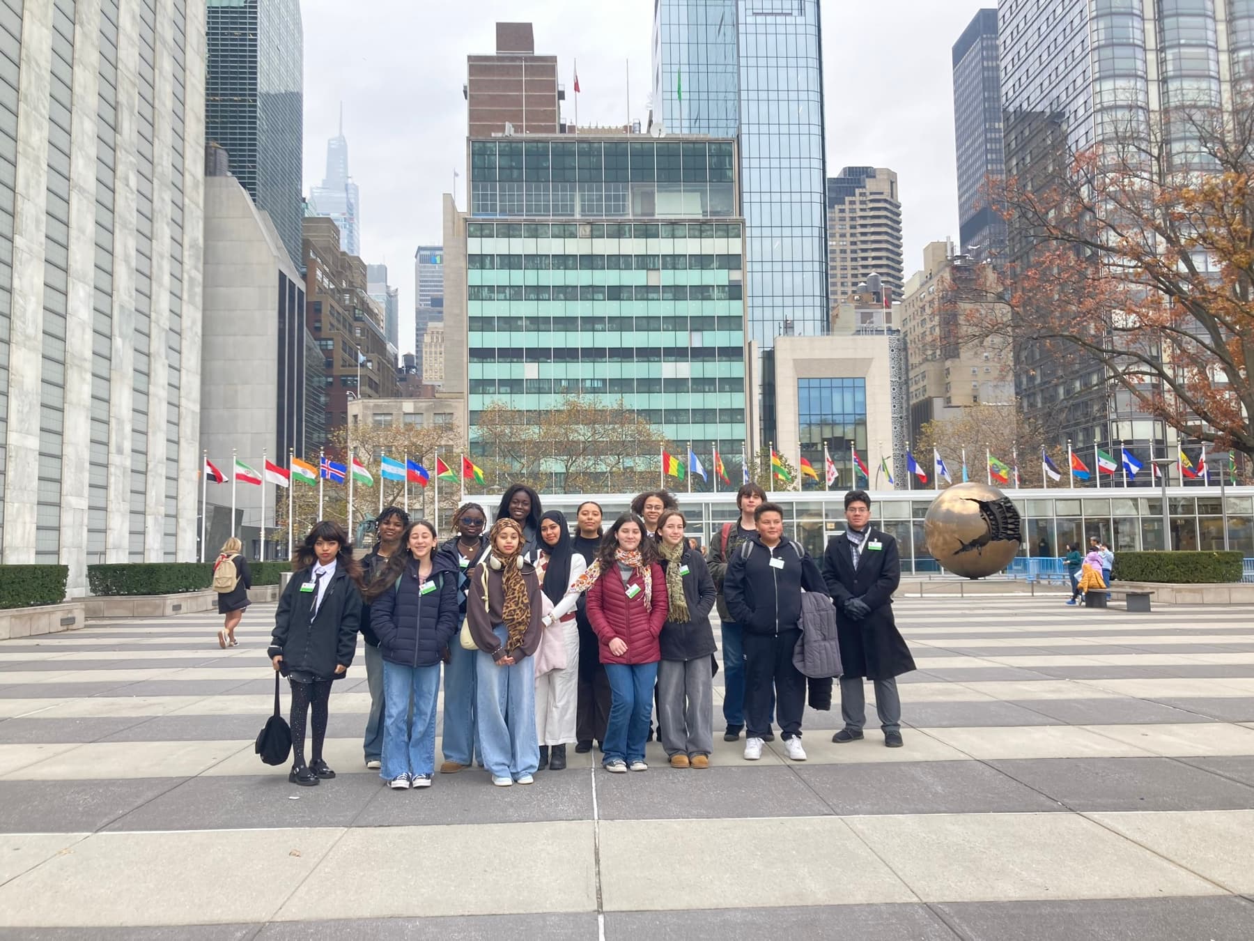 A group of high-school students wearing visitor badges standing in the plaza at United Nations Headquarters in New York, with the flags of the member states and the Sphere Within Sphere sculpture behind them.