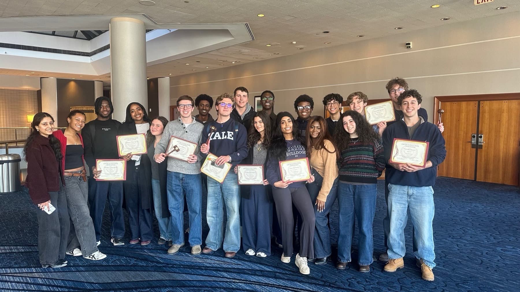 About twenty members of the Yale team standing together in a conference hotel, several holding framed award certificates and one holding a gavel.