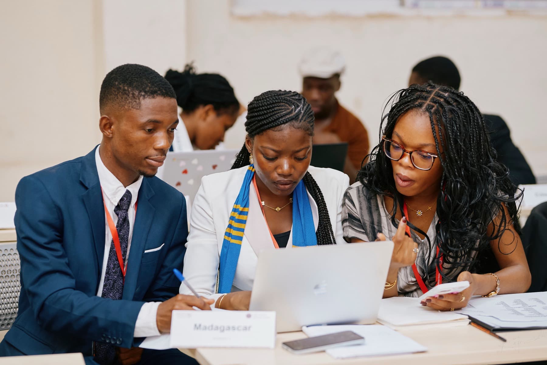 Three delegates leaning over a laptop together at a committee table, a paper placard reading Madagascar in front of them.