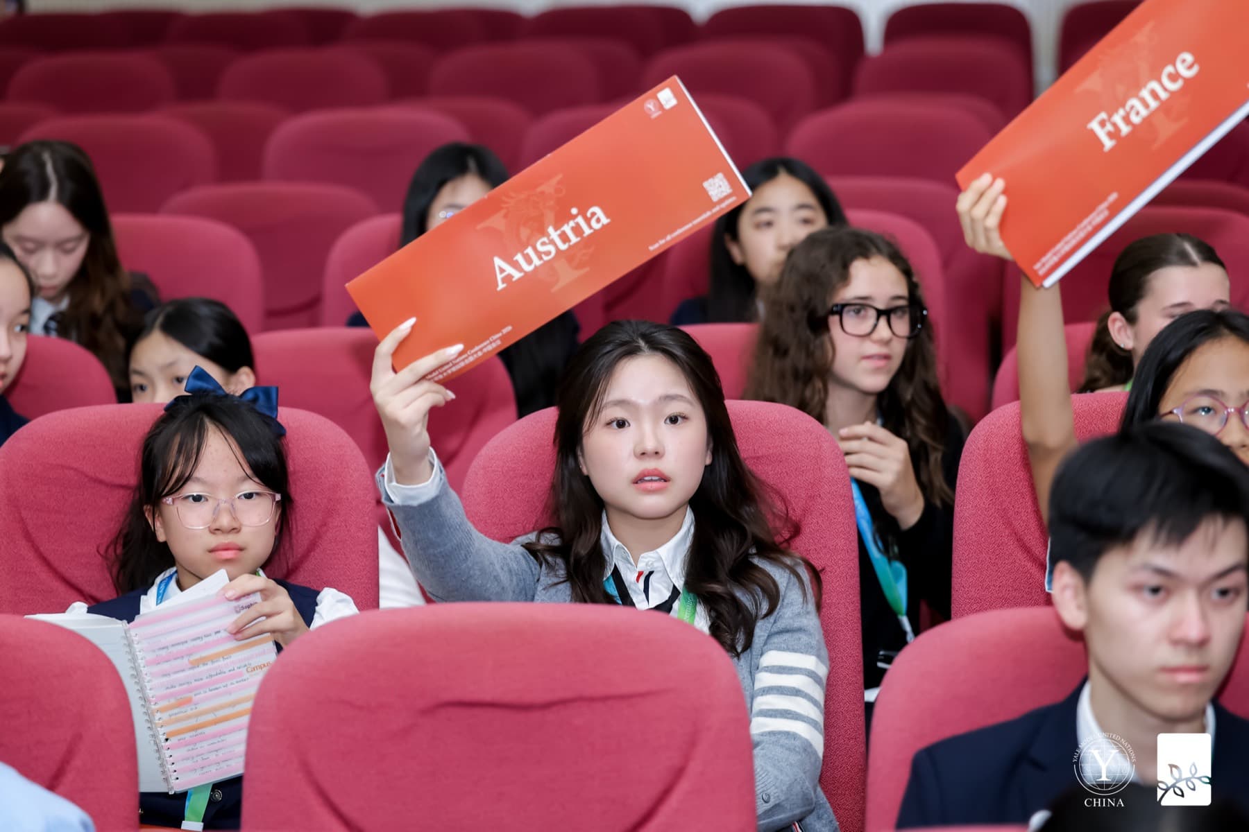 Delegates seated in a red auditorium, two of them holding up long orange placards reading Austria and France.