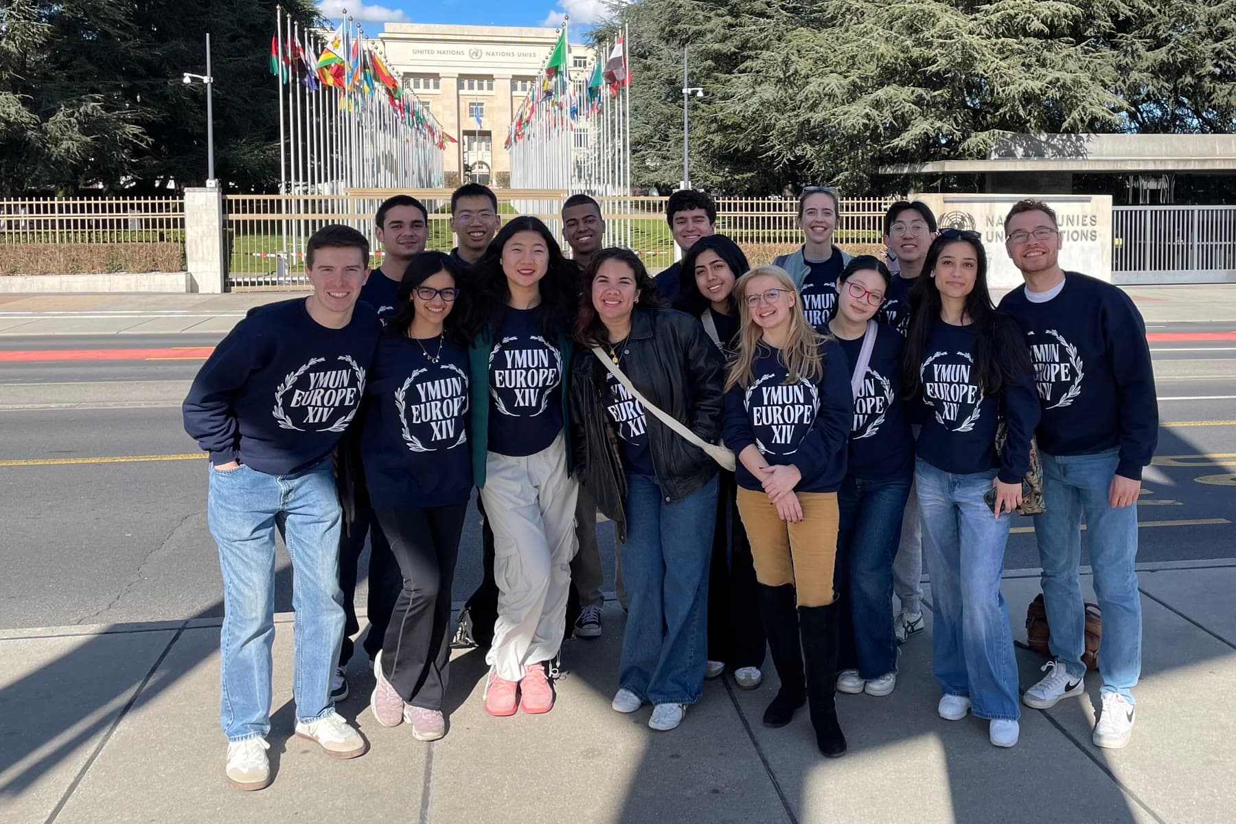 Around fifteen students in matching navy YMUN Europe sweatshirts standing on the pavement outside the Palais des Nations in Geneva, the flags of the member states behind them.