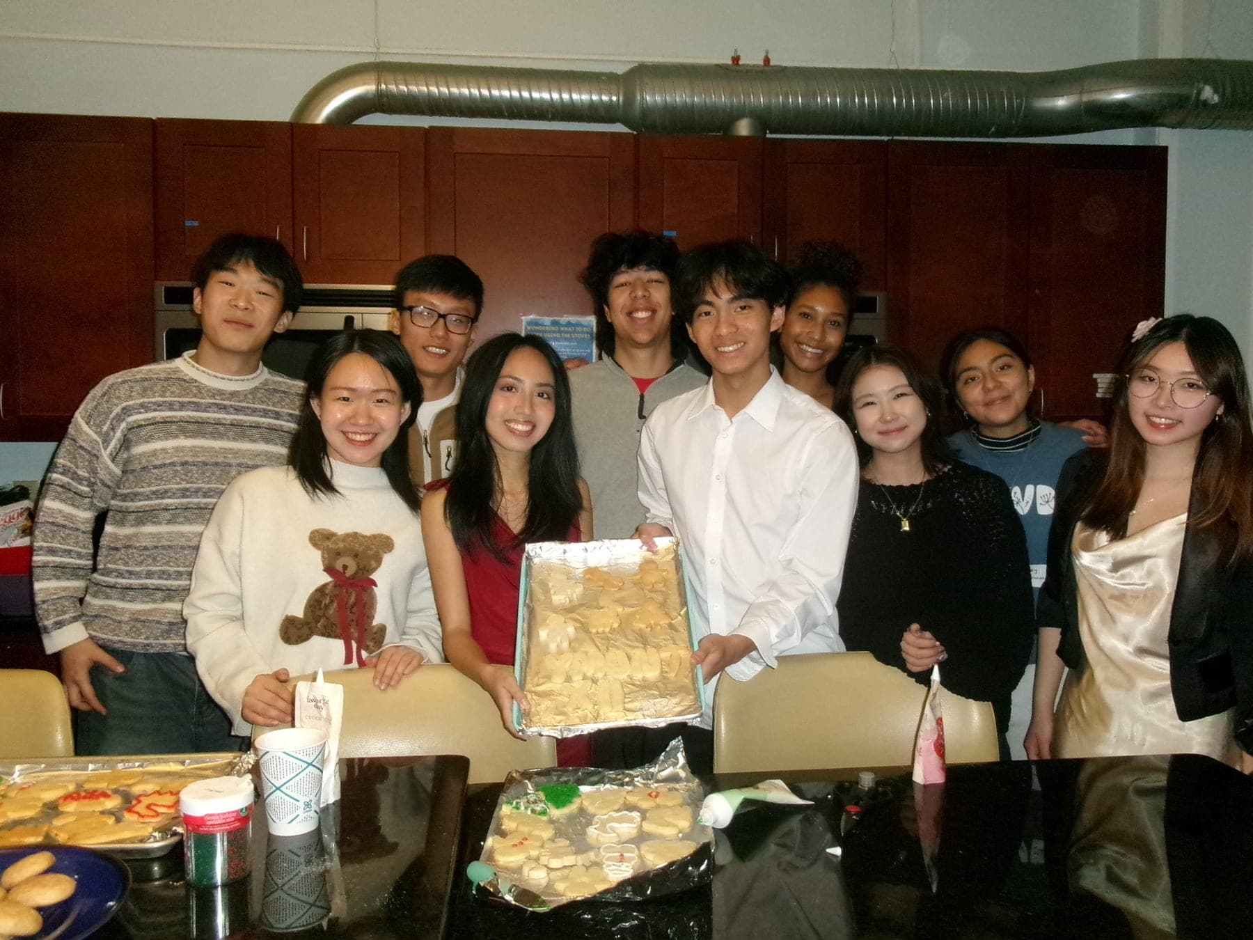 About a dozen students crowded together behind a kitchen counter covered in trays of iced sugar cookies, one of them holding up a baking sheet of gingerbread shapes.