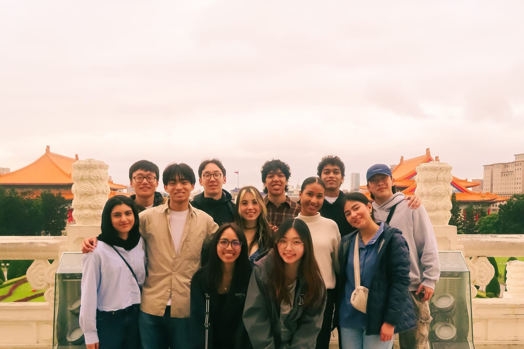 About a dozen students standing together on the terrace at Liberty Square in Taipei, the orange roofs of the National Theater and National Concert Hall behind them under a white sky.