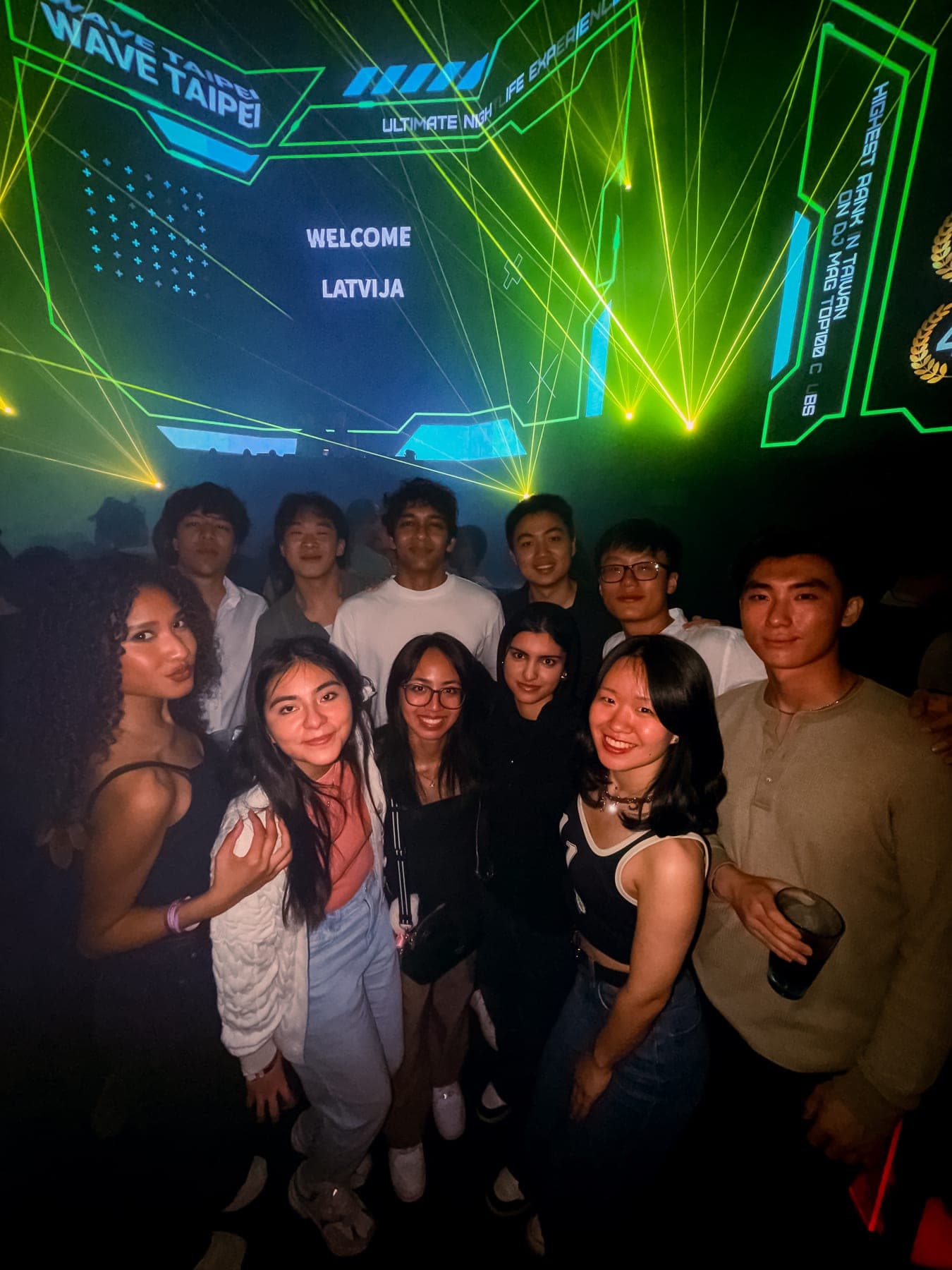Eleven students posing together on a dark club floor beneath green and yellow laser light, a screen reading Wave Taipei on the wall behind them.