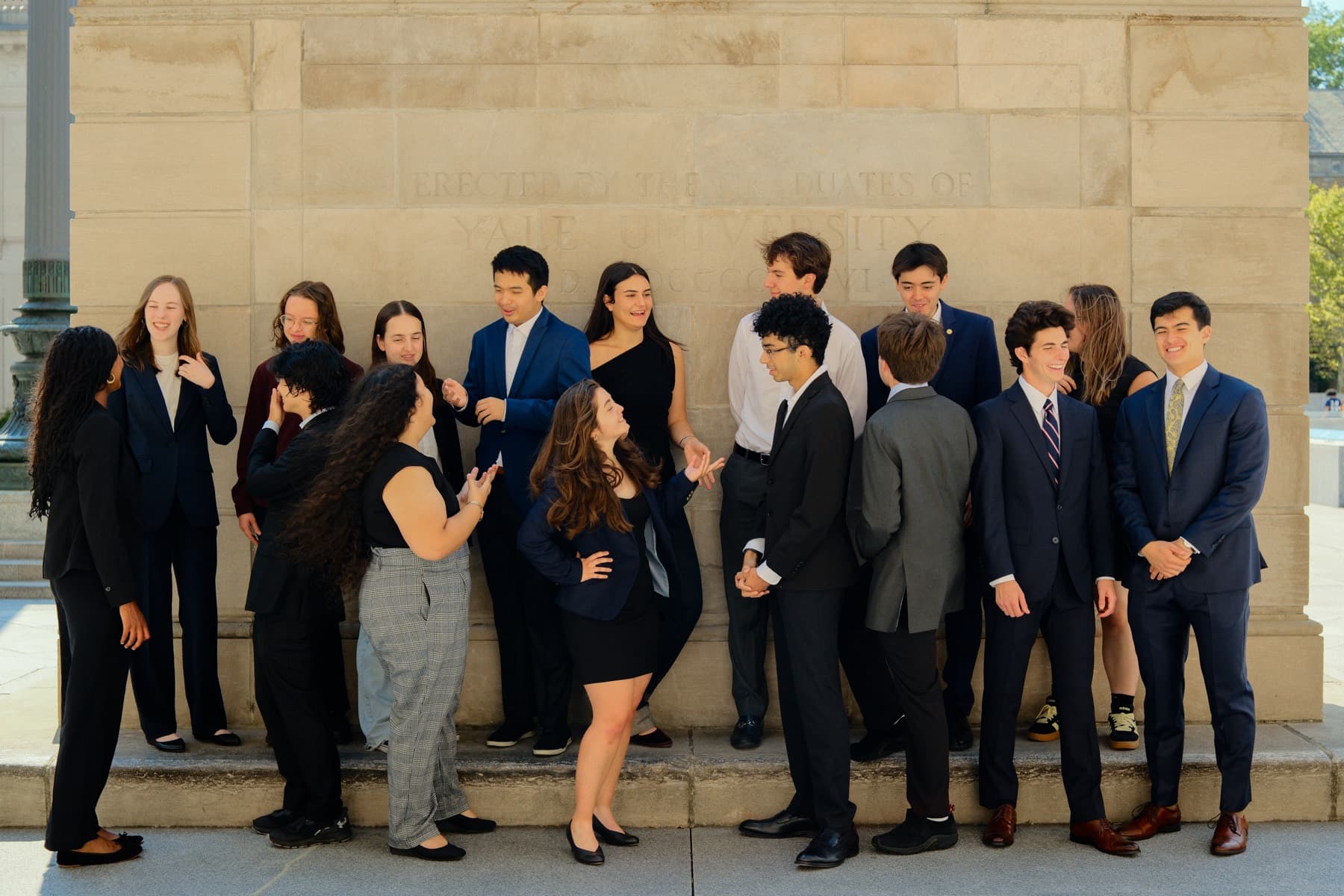 Around fifteen students standing in a line against a stone wall on Yale's campus, dressed formally.