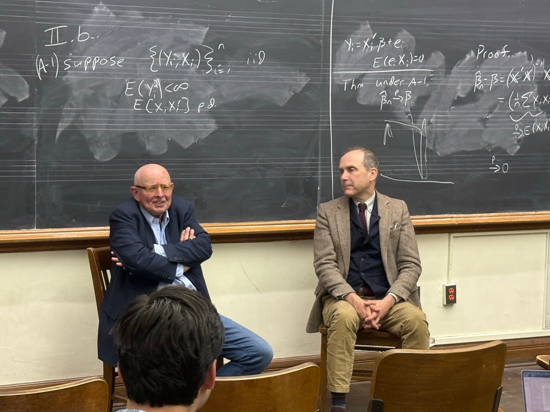 Two speakers sitting at the front of a Yale classroom in front of a blackboard covered in handwritten equations, talking to students seated in the rows below.